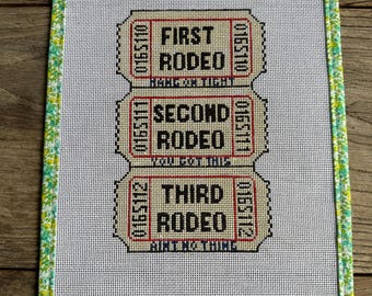 May include: Needlepoint art piece featuring three rodeo tickets with the text "First Rodeo", "Second Rodeo", and "Third Rodeo". The tickets are in red and tan, with black text. The piece is framed with a green floral border.