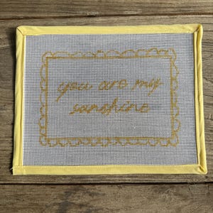 May include: A needlepoint canvas with the words "you are my sunshine" stitched in gold thread. The canvas is framed with a yellow fabric border. The background is a white and blue grid.