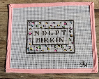 May include: A needlepoint canvas with a pink border and a white mesh center. The center features a brown rectangular design with the text "NDLPT BIRKIN" in black. Colorful, small, square designs surround the text.