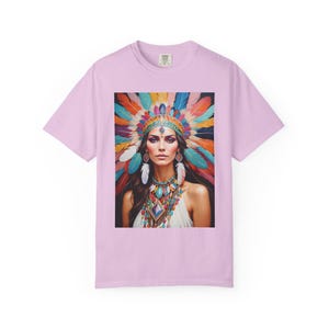 Könnte beinhalten: Hellrosa T-Shirt mit einem lebendigen Porträt einer Person, die mit einem farbenfrohen Federkopfschmuck und aufwendigem Schmuck geschmückt ist. Das Kunstwerk zeigt eine Frau mit langen dunklen Haaren, die ein weißes Oberteil und einen mehrfarbigen Kopfschmuck trägt.