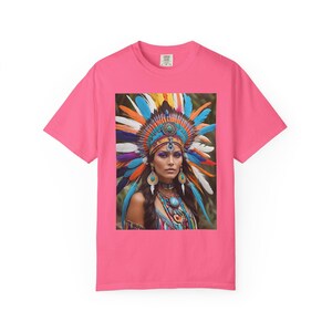 Könnte beinhalten: Ein pinkfarbenes T-Shirt mit einem Bild einer Person mit farbenfrohem Federkopfschmuck und türkisfarbenem Schmuck. Der Kopfschmuck zeigt Federn in Blau, Orange, Gelb und Lila. Das Bild ist auf dem T-Shirt abgebildet.