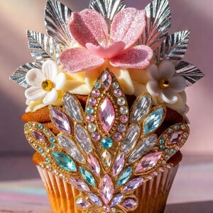 Könnte beinhalten: Ein dekorierter Cupcake mit einem Schmuckkronen-Design. Der Cupcake hat eine goldbraune Basis, gekrönt mit weißem Zuckerguss, rosa Blüten, silbernen Blättern und einer Gold- und Schmuckverzierung. Die Juwelen sind in Rosa-, Blau- und Klartönen gehalten.
