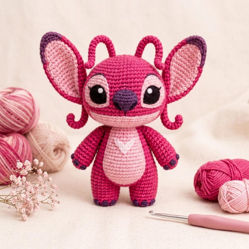 May include: A crochet amigurumi doll of a character with large ears, antennae, and a dark purple nose. The doll is primarily bright pink with a light pink belly patch and dark purple accents on the ears, nose, and feet, crafted with yarn.