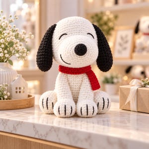 May include: A white crocheted dog figurine with long black ears, a black nose, and a closed-eye expression. The dog is wearing a bright red crocheted scarf around its neck. The paws feature black stitched details on a cream-coloured yarn body.