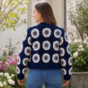 May include: A navy blue crochet sweater featuring a sunflower design. The sweater has a cropped fit and long sleeves. The sunflower pattern includes brown, white, and light blue details. The person in the image is wearing blue jeans.