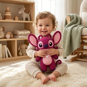 May include: A handmade crochet plush toy featuring a dark magenta and light pink alien character with large ears, a round head, and a small white heart detail on the chest. The toy has a textured yarn finish and a soft, stuffed construction.