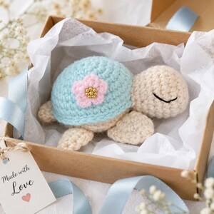May include: A crocheted turtle toy with a light blue shell featuring a pink flower accent with gold beads, resting inside a brown cardboard gift box lined with white tissue paper. A tag attached to the box reads Made with Love with a small heart.
