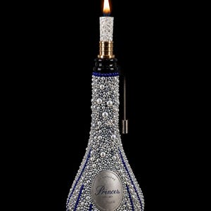 Può includere: Una lampada a bottiglia decorativa composta da una bottiglia di champagne Princes de Venoge ricoperta di perle argentate e bianche con accenti di perline blu verticali. Il collo è sormontato da una candela bianca a motivo floreale accesa.