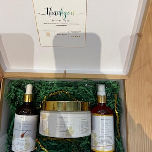Op de afbeelding: Een geschenkdoos met drie schoonheidsproducten: een bruine glazen fles met een druppelaar, een witte pot met een gouden deksel en een rode spuitfles. De producten zijn voorzien van het merk "Belle Q'iss". De doos is gevuld met groen papier.