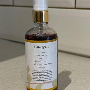 Op de afbeelding: Een heldere glazen fles van Belle.Q'iss Organic Aloe Vera and Rose Water Hydration Hair Mist. De fles heeft een gouden dop en een witte sproeikop. Het etiket bevat tekst over de ingrediënten en voordelen van het product. De fles bevat 100 ml product.
