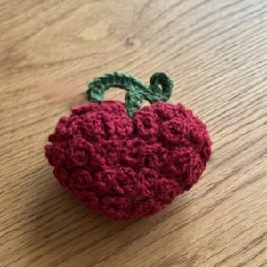 Op de afbeelding: Een handgemaakte gehaakte aardbei met een groene steel. De aardbei heeft een dieprode kleur en een getextureerd oppervlak. De steel is heldergroen. De aardbei ligt op een houten oppervlak.