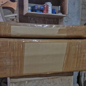 May include: Two rectangular wooden objects stacked vertically, each wrapped in clear plastic film and secured with wide tan adhesive tape. The surfaces are smooth and light brown, showing the natural grain of the wood beneath the protective packaging.