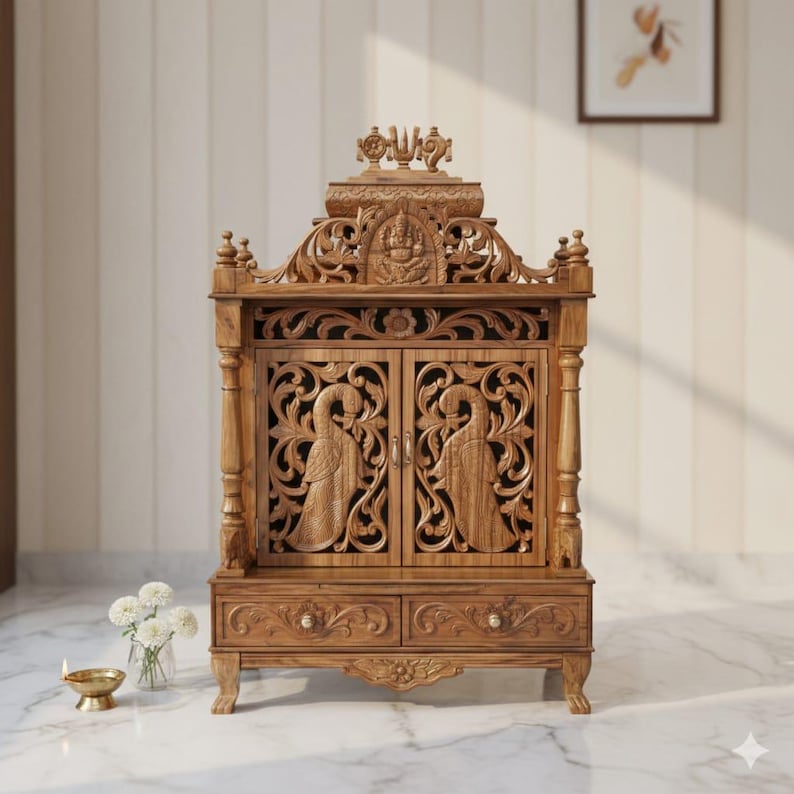 May include: A wooden home temple featuring intricate carvings of two peacocks on the cabinet doors, a central deity figure at the top, and two drawers at the base. The structure is supported by four carved legs and displays a natural wood grain finish.