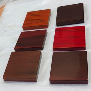 May include: Six square wooden blocks arranged in two columns of three. Each block features a distinct wood grain pattern and a polished finish, displaying a variety of warm tones including light orange, deep red, and dark brown wood shades.