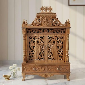 May include: A brown wooden home temple featuring intricate hand-carved details, including two doors with peacock motifs, a central deity carving at the top, and two lower drawers with floral patterns, supported by four carved legs.