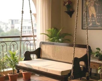 May include: A dark wood swing with a woven back and a beige cushion hangs from the ceiling. The swing is suspended by chains and is on a balcony with potted plants and a view of a city.