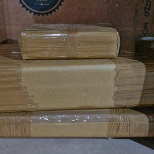 May include: Three rectangular packages wrapped in plain brown paper and secured with clear adhesive tape. The items are stacked in a pyramid formation, with one smaller package resting on top of two larger, flatter packages of equal width.