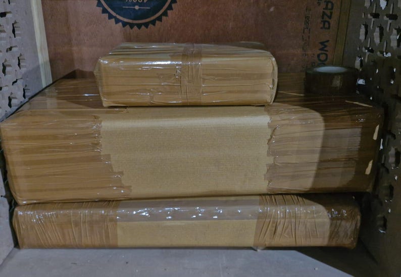 May include: Three rectangular packages wrapped in plain brown paper and secured with clear adhesive tape. The items are stacked in a pyramid formation, with one smaller package resting on top of two larger, flatter packages of equal width.