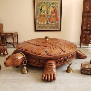 May include: A large, hand-carved wooden turtle-shaped table with a shell featuring a lotus petal design. The turtle has a brass lotus finial on top, brass bells hanging from its neck and front legs, and a detailed carved border around the shell.