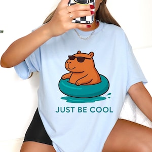 May include: Pale blue t-shirt with a cartoon capybara wearing sunglasses in a teal inflatable ring. The text "JUST BE COOL" is printed below the graphic. The shirt has a loose fit.