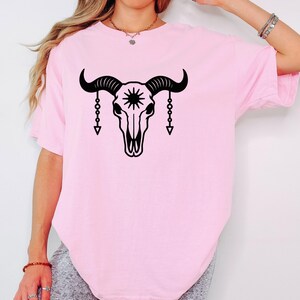 Op de afbeelding: Lichtroze T-shirt met een zwarte afbeelding van een stierschedel met een zonnester en hangende kettingaccenten. Het shirt heeft een comfortabele, casual stijl.