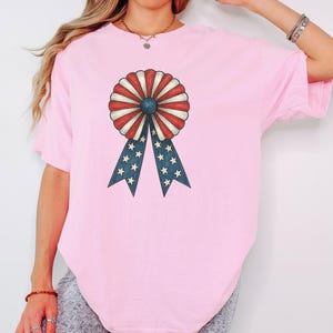 Könnte beinhalten: Hellrosa T-Shirt mit einem patriotischen Rosetten-Design. Die Rosette hat rot-weiß gestreifte Blütenblätter, eine blaue Mitte und blaue Bänder mit weißen Sternen. Das Shirt ist ein bequemer, lässiger Stil.