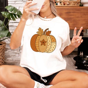 May include: White t-shirt with a patchwork pumpkin design in orange, brown, and tan. The pumpkin has a star and leaf accents. The shirt is a casual style.