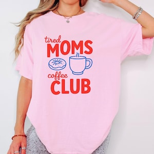 Op de afbeelding: Een lichtroze t-shirt met de tekst "tired MOMS coffee CLUB" in rood en blauw. Het ontwerp bevat een blauwe donut en een blauwe koffiekop met stoom.