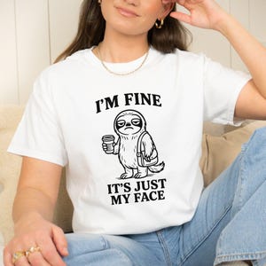 May include: White t-shirt with the text "I'M FINE IT'S JUST MY FACE" and a black and white illustration of a sloth holding a coffee cup and a bag. The sloth has a grumpy expression.