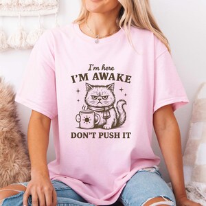 Può includere: T-shirt rosa chiaro con un'illustrazione di un gatto scontroso che tiene una tazza di caffè con un disegno di sole. Il testo recita "I'm here I'm awake DON'T PUSH IT" in marrone. La maglietta è a girocollo e maniche corte.