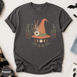 May include: A charcoal gray short-sleeve t-shirt featuring a graphic of an orange witch hat adorned with flowers and wheat. Below the hat are moon phases and botanical sprigs. The shirt has a crew neckline and a relaxed, casual fit.