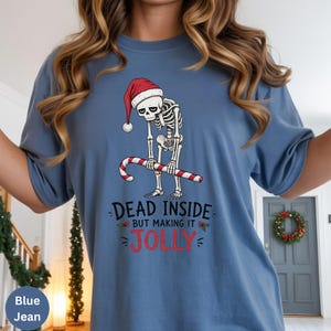 May include: A blue jean colored t-shirt featuring a graphic of a skeleton wearing a red and white Santa hat, holding a red and white candy cane. Below the skeleton, the text reads: DEAD INSIDE BUT MAKING IT JOLLY, with small holly accents.