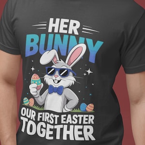 Op de afbeelding: Zwart T-shirt met een afbeelding van een cartoonkonijn met een zonnebril en een vlinderdas, met een versierd paasei. Tekst luidt "HER BUNNY OUR FIRST EASTER TOGETHER" in wit en blauw.