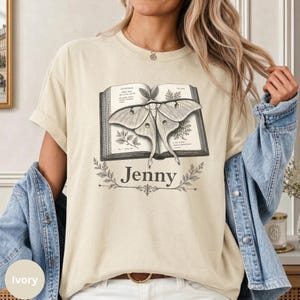 May include: An ivory short-sleeve t-shirt featuring a graphic of an open book with a moth illustration on the pages. The name Jenny is printed in black serif font below the book, framed by decorative botanical line art elements.