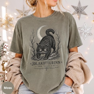 Puede incluir: Una camiseta de manga corta color verde musgo con una ilustración gráfica negra de un gato bajo una luna creciente y árboles desnudos. El texto dice JOLAKOTTURINN THE YULE CAT. La camiseta tiene cuello redondo y un aspecto desgastado.