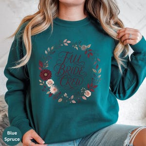 May include: A dark teal crewneck sweatshirt featuring a floral wreath graphic in shades of burgundy, cream, and muted green. The text "FALL BRIDE CREW" is printed in a dark red cursive script in the center of the wreath design.
