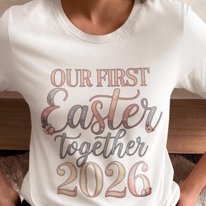 Puede incluir: Camiseta blanca con el texto "OUR FIRST Easter together 2026" en tonos pastel. El diseño presenta elementos florales y huevos, ideal para celebraciones de Pascua.