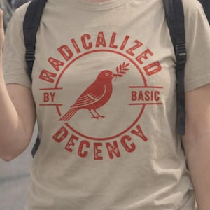 Peut inclure: Un t-shirt beige avec un motif rouge. Le motif représente un oiseau tenant une branche dans un cercle. Le texte autour du cercle se lit "RADICALIZED BY BASIC DECENCY".