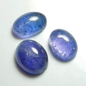 Puede incluir: Tres piedras preciosas ovaladas pulidas en tonos azules y morados. Las piedras tienen una superficie lisa y brillante y patrones internos variables. Las piedras preciosas se muestran sobre un fondo blanco.