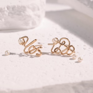 May include: Gold-toned wire earrings spelling out "Yes" and "Love", each adorned with a small pearl. The earrings are displayed on a textured white surface, with additional pearls scattered nearby.