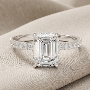 May include: A silver engagement ring with a large, rectangular emerald-cut diamond as the centerpiece. The band is encrusted with smaller diamonds, adding sparkle. The ring is set against a neutral-toned fabric background.