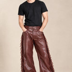 Könnte beinhalten: Ein schlichtes schwarzes T-Shirt mit kurzen Ärmeln kombiniert mit einer weit geschnittenen dunkelbraunen Lederhose. Die Hose ist an den äußeren Seitennähten beider Beine mit langen, dünnen Lederfransen versehen und hat einen klassischen Knopfverschluss.