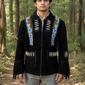 Könnte beinhalten: Eine schwarze Wildlederjacke mit langen Fransen an Brust und Ärmeln, akzentuiert durch weiße Knochenperlen und blaue Perlenpaneele mit Adlerdarstellungen. Die Jacke verfügt über einen Frontreißverschluss und einen runden Aufnäher am linken Ärmel.