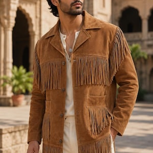 Könnte beinhalten: Eine hellbraune Wildlederjacke mit langen Fransen an Brust, Schultern und den unteren Taschen. Die Jacke hat einen Kragen, eine Knopfleiste auf der Vorderseite und lange Ärmel. Sie wird über einem cremefarbenen Hemd und einer hellen Hose getragen.