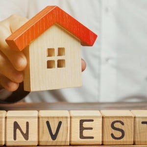 May include: A wooden toy house with an orange roof is held above wooden blocks spelling out the word "INVEST". The house has four square windows. The blocks are on a wooden surface.