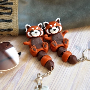 Peut inclure: Deux porte-clés représentant des pandas roux. Chaque porte-clé présente une figurine de panda roux avec un visage blanc, un corps marron et une queue segmentée. Les porte-clés sont attachés à une chaîne et un anneau argentés. Le fond comprend des lunettes de soleil et un sac en toile.