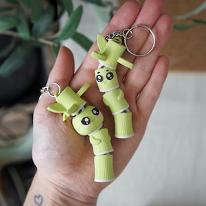 Peut inclure: Deux porte-clés en forme de bambou vert clair avec des corps cylindriques segmentés et des surfaces texturées. Chaque porte-clé possède une petite tête de personnage avec de grands yeux noirs, des reflets blancs et des joues roses, fixé à un anneau en métal argenté.