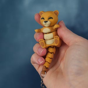 Op de afbeelding: Een sleutelhanger van een lachende, bruine leeuwenwelp met een gesegmenteerd lichaam en staart. De welp heeft een crèmekleurige buik en poten en is bevestigd aan een zilveren sleutelring. De achtergrond is een wazig, donkerblauw.