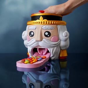 Puede incluir: Un dispensador de caramelos con forma de cascanueces caprichoso, con una cara detallada, barba blanca y sombrero negro y dorado. La boca del dispensador contiene una bandeja de caramelos coloridos.