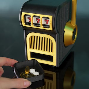 May include: A black and gold slot machine pill dispenser featuring three reels displaying the number seven. The device includes a side lever and a front dispensing tray. A small black square tray contains two white round pills and two yellow oval capsules.